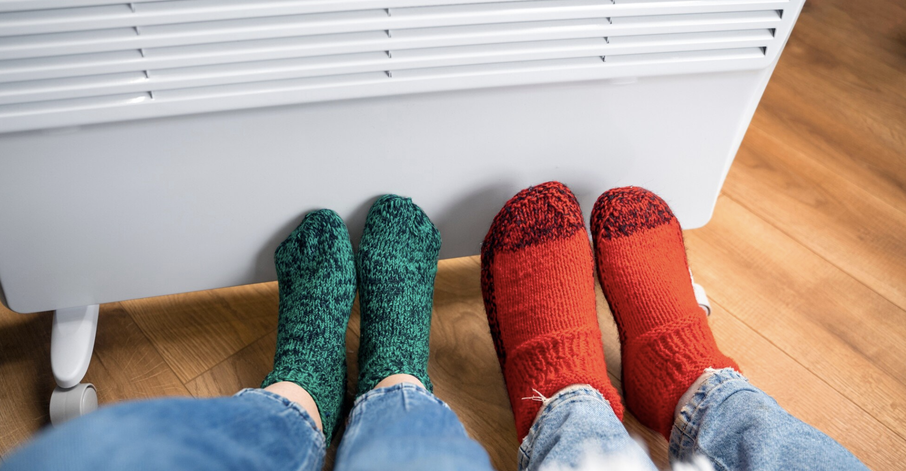 Deux personnes en chaussettes colorées, l'une portant des chaussettes vertes et l'autre des chaussettes rouges, réchauffent leurs pieds devant un radiateur blanc sur un sol en bois.